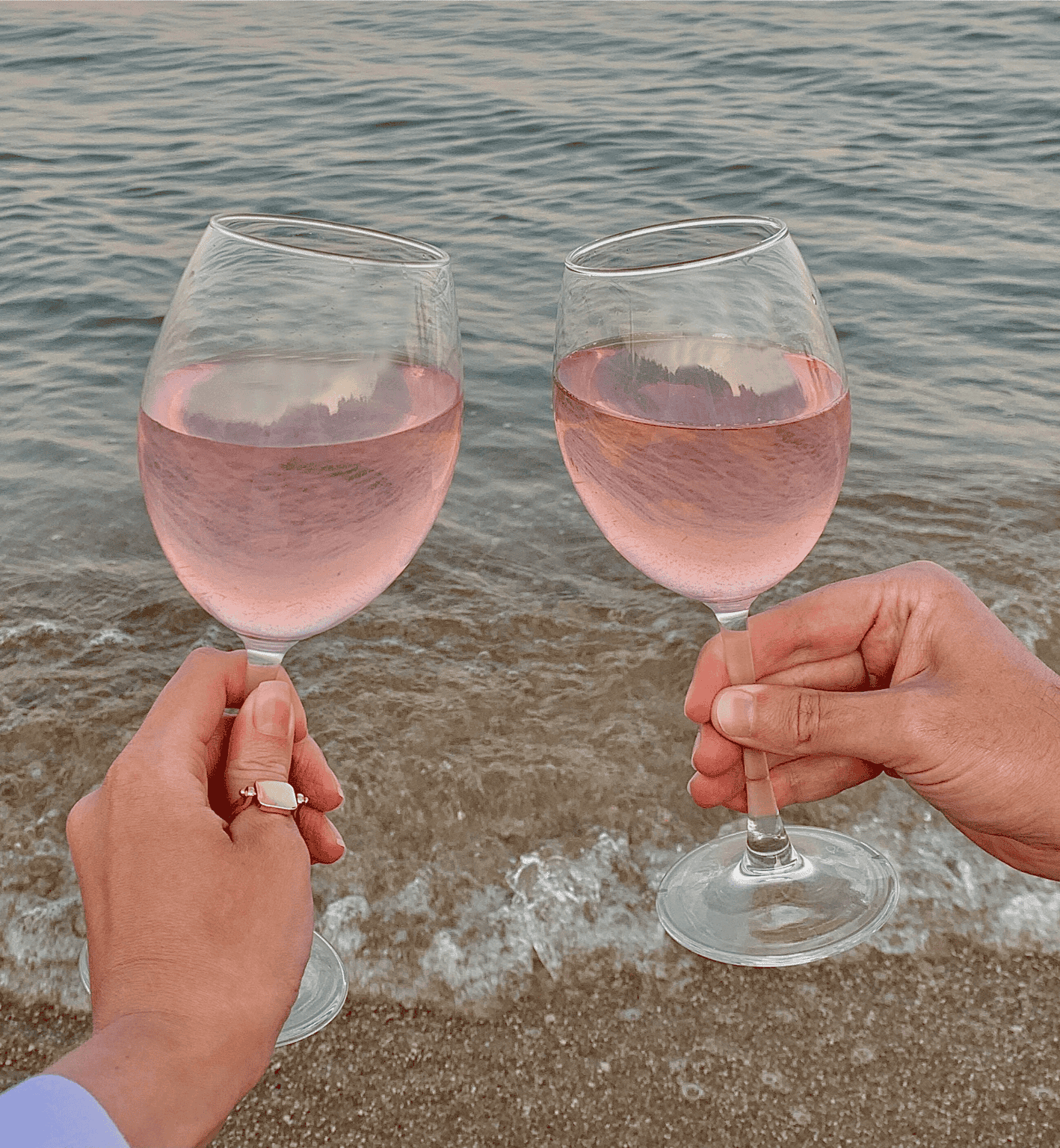 Two glasses of ros wine clinking together at the beach with ocean waves in the background, celebrating a seaside moment.